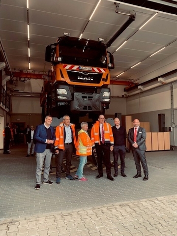 Leitung und Mitarbeiter stehen vor einem Fahrzeug der Straßenmeisterei, das in einer Halle hochgebockt ist. Es sieht so aus als schwebe das Fahrzeug über den Menschen