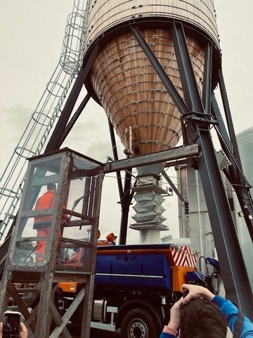 Das Foto zeigt einen riesigen Trichter aus dem Sand in ein dadrunter stehendes Fahrzeug verladen wird. Ein Mitarbeiter steht auf einer geschützten Kanzel und bedient das Konstrukt per Fernbedienung.