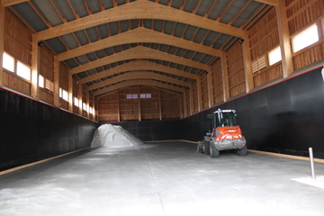 Das Foto zeigt eine große Lagerhalle. Es ist eine Holzkonstruktion mit Fenstern. Dort hinten links in der Ecke ist ein Haufen Sand, rechts in der Mitte steht ein Bagger.