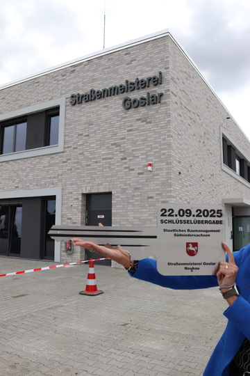 das neue Gebäude mit großen Buchstaben "Straßenmeisterei Goslar" ist zu sehen. Es ist noch ein Absperrband und ein Poller auf dem Gelände. Davor steht jemand mit dem Schild: 22.09.2025 Schlüsselübergabe SBN/Straßenmeisterei Goslar Neubau