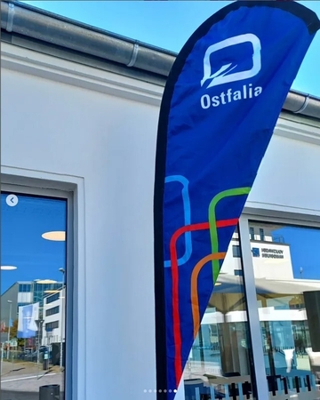 Fahnenaufsteller mit dem Zeichen der Ostfalia Hochschule und der Aufschrift Ostfalia. Im Hintergrund spiegelt sich in den Fenstern die Umgebung.
