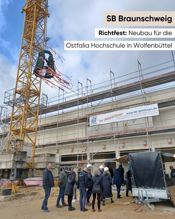 der Richtkranz für den Bau ist zu sehen. Er hängt am Kran. Unten werden mehrere Menschen durch die Baustelle geführt.