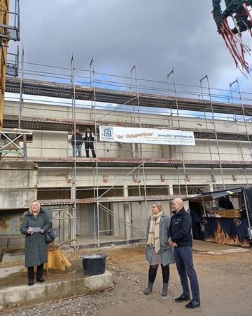 eine Frau steht etwas erhöht und trägt was vor. Man sieht eine Frau und einen Mann als Zuhörer. Der Rohbau ist im Hintergrund mit Gerüst zu sehen.