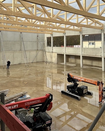 Das Foto zeigt große Baumaschinen im Rohbau des Gebäudes. Im Hintergrund steht ein Arbeiter. Die Dachkonstruktion aus Holz ist zu sehen und es steht Regenwasser auf dem Boden