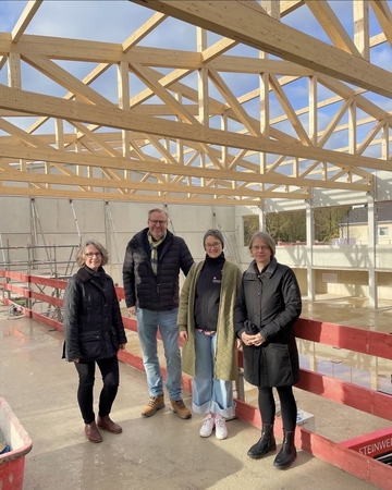 drei Frauen und ein Mann stehen im Rohbau ganz oben an einem provisorischen Geländer. Oben sieht man Teile der Holzdachkonstruktion im Rohbau