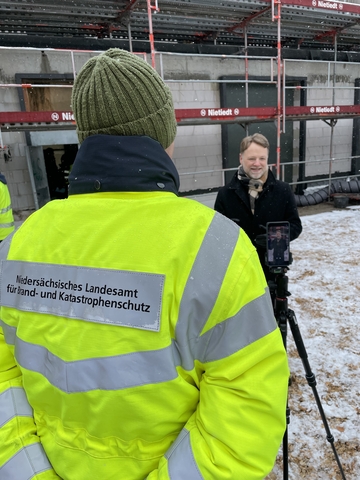Minister Heere steht vor der Kamera und mit dem Rücken zum Rohbau. Im Vordergrund steht ein Mitarbeiter des Niedersächsischen Landesamt für Brand- und Katastrophenschutz