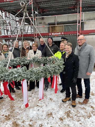 Finanzminister Heere, Innenministerin Behrends und weitere Menschen beim Richtfest Loy. Der Richtkranz hängt vor den Zuschauern und vor dem eingerüstetem Rohbau