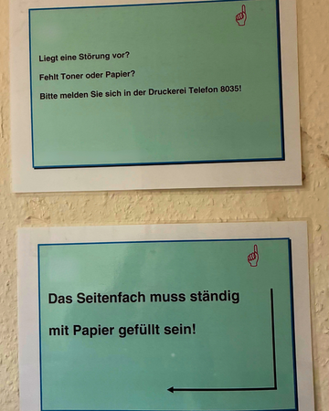 Zwei Schilder an einer Wand. Oben steht "Liegt eine Störung vor? Fehlt Toner oder Papier? Bitte melden Sie sich in der Druckerei 8035"; unten steht "Das Seitenfach muss ständig gefüllt sein"