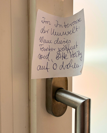 handgeschriebener Zettel an einem Griff vom Fenster oder an der Tür mit der Aufschrift: Im Interesse der Umwelt: Wenn dieses Fenster geöffnet wird, bitte Heizung auf 0 drehen.
