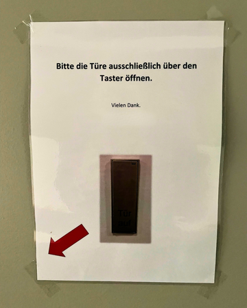 auf einem mit an den Ecken mit Tesa geklebten Blatt Papier ist das Abbild eines Türtasters zu erkennen mit einem Richtungspfeil, da drüber steht "Bitte die Türe ausschließlich über den Taster öffnen. Vielen Dank".