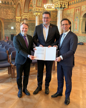 von links nach rechts stehen Finanzminister Heere, Oberbürgermeister Kornblum und Wissenschafts- und Kulturminister Mohrs und halten im Saal den unterzeichneten Vertrag in die Kamera