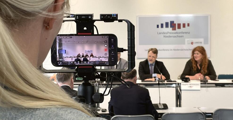 Finanzminister Heere bei eine Pressekonferenz durch eine Kamera einer Medienvertreterin