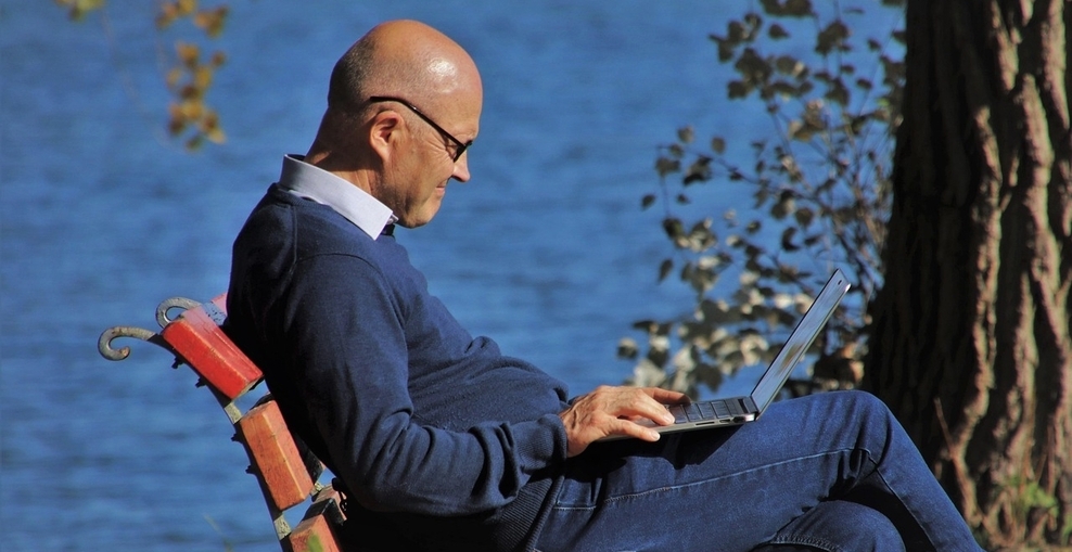 Älterer Mann sitzt auf Bank vor einem See. Auf seinem Schoß liegt ein aufgeklappter Laptop.